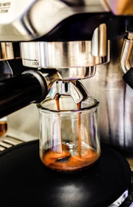 Espresso coffee flows smoothly from a metal espresso machine into a glass cup, highlighting the rich, golden-brown crema forming at the top of the liquid.