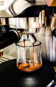 Espresso coffee flows smoothly from a metal espresso machine into a glass cup, highlighting the rich, golden-brown crema forming at the top of the liquid.
