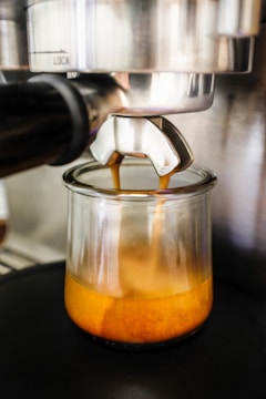 A close-up view of an espresso machine pouring fresh espresso into a small glass cup. The rich crema is visible as the coffee fills the container. The metallic finish of the machine contrasts with the brown hues of the espresso.
