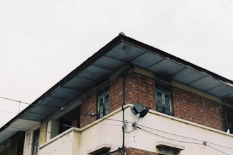 A two-story building with exposed brickwork on the upper floor and concrete walls below. The structure has a flat metal roof with visible electrical wiring along the exterior, and there are two wooden windows on the upper floor.