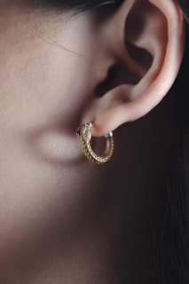 A close-up of a person's ear adorned with a hoop earring shaped like a snake. The earring is golden with a detailed, textured design resembling scales. The ear's outline is clearly visible, and the lighting highlights the smooth skin and the earring's metallic sheen.
