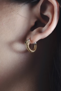 A close-up of a person's ear adorned with a hoop earring shaped like a snake. The earring is golden with a detailed, textured design resembling scales. The ear's outline is clearly visible, and the lighting highlights the smooth skin and the earring's metallic sheen.