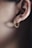 A close-up of a person's ear adorned with a hoop earring shaped like a snake. The earring is golden with a detailed, textured design resembling scales. The ear's outline is clearly visible, and the lighting highlights the smooth skin and the earring's metallic sheen.