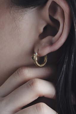 A close-up of an ear adorned with a gold, textured hoop earring resembling a coiled snake. Fingers gently touch the area near the earring, emphasizing the jewelry's intricate details and the texture of the skin.