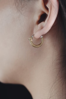 A close-up view of a person's ear adorned with a gold hoop earring. The earring features a textured design with a small, intricate pattern. The skin tone is neutral, and fine strands of hair are visible along the edge.