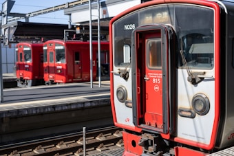A vibrant red commuter train is stationed on the tracks at a modern railway platform. The train is marked with the number 815 and belongs to the Kyushu Railway Company. Additional red train cars are visible in the background, highlighting a busy and organized railway setting. The platform is equipped with overhead structures and signs, indicating a well-maintained and efficient railway station.