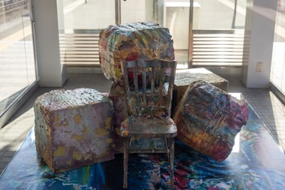 A wooden chair is positioned among several large, abstract, colorful stone-like blocks. The blocks are intricately painted with vibrant hues and textures, giving them a marbled appearance. The setting is indoors with large windows allowing natural light to illuminate the objects, enhancing their colors.