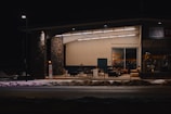 A building with a stone and wood facade is depicted under dim street lighting. The interior is illuminated, revealing various furniture pieces and a sign that reads 'Idaho Youth Ranch'. Snow is present on the ground in front of the building.
