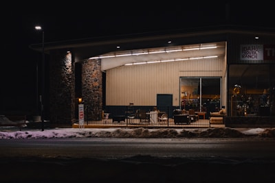A building with a stone and wood facade is depicted under dim street lighting. The interior is illuminated, revealing various furniture pieces and a sign that reads 'Idaho Youth Ranch'. Snow is present on the ground in front of the building.