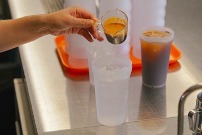 A hand holds a small cup of espresso over a larger cup filled with ice. Several containers and a tray are visible on a metallic countertop. An iced coffee is placed on the surface nearby.