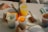 Four drinks in plastic cups are arranged on a table, each containing different types of beverages like iced coffee, lemonade with herbs, and an orange drink. There is a slice of bread on a red plate and a small cake on a white plate, accompanied by a fork. An open book is visible at the bottom of the image.