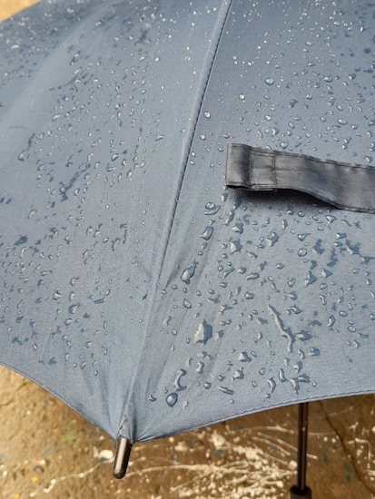 A close-up view of a blue umbrella covered in water droplets, suggesting recent rainfall. The image highlights the texture of the fabric and the even distribution of droplets across the surface.
