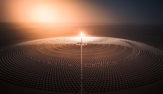 A vast array of solar panels is arranged in concentric circles around a central tower with a bright light, likely depicting a solar power plant. The setting appears to be a desert with a hazy sunrise or sunset casting an orange glow across the landscape.