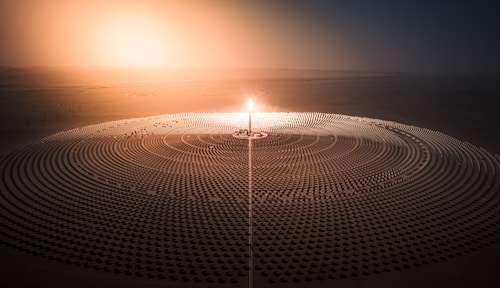 A vast array of solar panels is arranged in concentric circles around a central tower with a bright light, likely depicting a solar power plant. The setting appears to be a desert with a hazy sunrise or sunset casting an orange glow across the landscape.