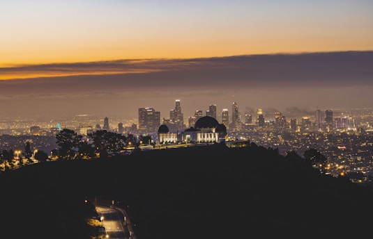 A cityscape at sunset with a large observatory in the foreground, silhouetted against the city skyline. The sky is a gradient of orange and blue hues, while city lights twinkle in the distance.