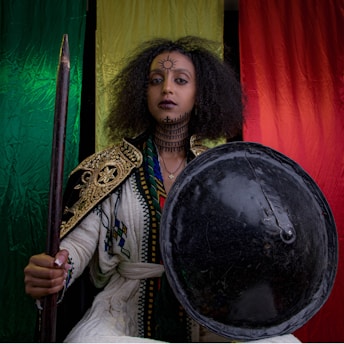 A person wearing traditional attire with intricate, gold-embroidered details stands confidently holding a spear and shield. The individual has distinctive facial and neck tattoos, and their hair is styled in a voluminous afro. Behind them are vibrant green, yellow, and red vertical banners.