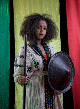 A person with curly hair, adorned with traditional facial markings, holds a spear and shield against a backdrop of vertical tricolors: green, yellow, and red. They wear a robe with intricate patterns.