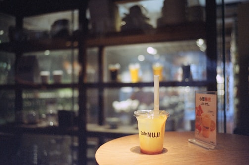 A plastic cup with a straw labeled 'Café MUJI' is positioned on a wooden table. Nearby, there is a small upright menu stand with images and text. The background features a dimly lit display with glassware and soft reflections.