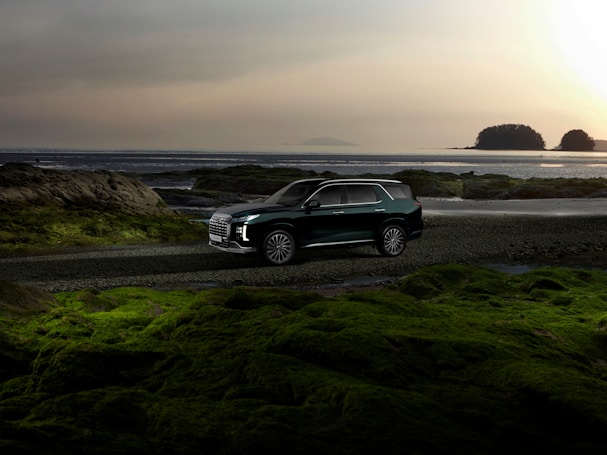 A dark-colored SUV is parked on a coastal road with a scenic backdrop of the ocean and distant islands. The sky is cloudy, adding a serene and calm ambiance to the scene. Lush green moss covers the rocky surfaces in the foreground, contrasting with the vehicle's sleek, modern design.
