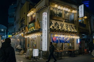 A traditional Japanese street scene at night featuring a cozy, warmly lit gyoza restaurant adorned with hanging lanterns and signs in Japanese. People are walking past, adding life to the setting. The building has a nostalgic and inviting ambiance.