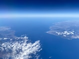 An aerial view of a coastal landscape featuring two landmasses separated by a large body of water. The land is marked by uneven terrain and scattered clouds, with a clear horizon line separating the deep blue sea from the sky.