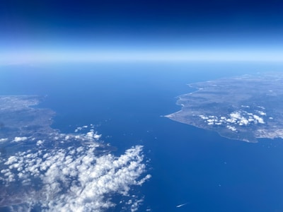 An aerial view of a coastal landscape featuring two landmasses separated by a large body of water. The land is marked by uneven terrain and scattered clouds, with a clear horizon line separating the deep blue sea from the sky.