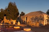 A serene outdoor setting features large canvas tents surrounded by tall trees. The tents have wooden chairs in front, and the area is decorated with traditional woven rugs and cushions. Delicately crafted lanterns are placed on the ground, and the scene is bathed in warm, golden sunlight, suggesting a sunrise or sunset.