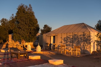 A serene outdoor setting features large canvas tents surrounded by tall trees. The tents have wooden chairs in front, and the area is decorated with traditional woven rugs and cushions. Delicately crafted lanterns are placed on the ground, and the scene is bathed in warm, golden sunlight, suggesting a sunrise or sunset.