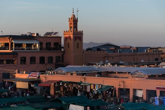 A bustling marketplace with numerous stalls covered by green tarps. A large clock tower is visible in the background along with several two-story buildings. The scene is illuminated by warm sunlight, suggesting it is either early morning or late afternoon. People mill about the market area, and mountains can be seen in the distance.