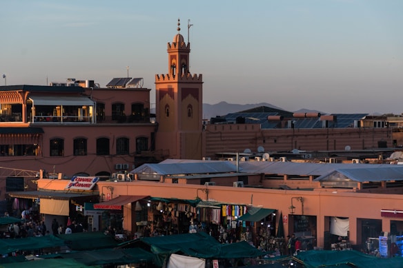 A bustling marketplace with numerous stalls covered by green tarps. A large clock tower is visible in the background along with several two-story buildings. The scene is illuminated by warm sunlight, suggesting it is either early morning or late afternoon. People mill about the market area, and mountains can be seen in the distance.