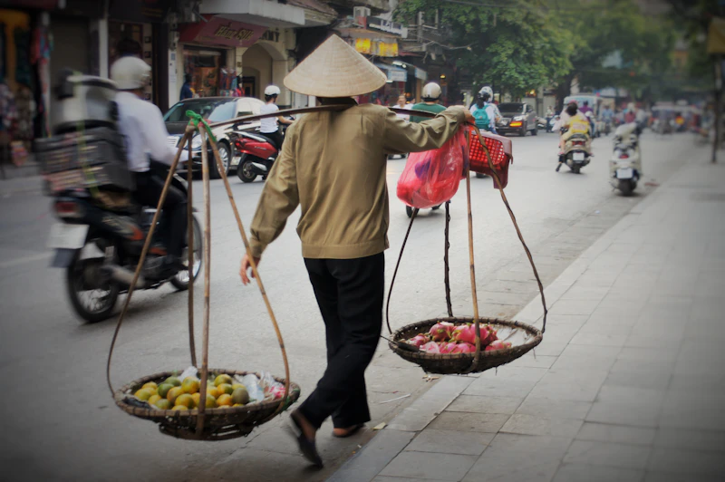 A vendor in a conical hat carrying two baskets of fruit