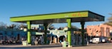 A vibrant green gas station with multiple fuel pumps is situated in a bright area surrounded by motorcycles, a van, and a distinct architectural building with intricate patterns. There are palm trees and flags in the background, adding to the lively and colorful ambiance.