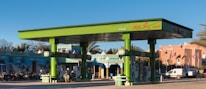 A vibrant green gas station with multiple fuel pumps is situated in a bright area surrounded by motorcycles, a van, and a distinct architectural building with intricate patterns. There are palm trees and flags in the background, adding to the lively and colorful ambiance.