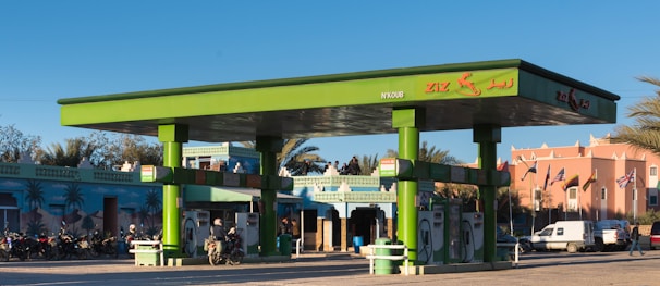 A vibrant green gas station with multiple fuel pumps is situated in a bright area surrounded by motorcycles, a van, and a distinct architectural building with intricate patterns. There are palm trees and flags in the background, adding to the lively and colorful ambiance.
