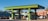 A vibrant green gas station with multiple fuel pumps is situated in a bright area surrounded by motorcycles, a van, and a distinct architectural building with intricate patterns. There are palm trees and flags in the background, adding to the lively and colorful ambiance.