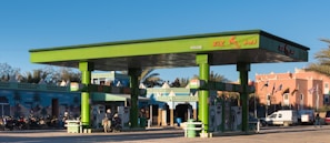 A vibrant green gas station with multiple fuel pumps is situated in a bright area surrounded by motorcycles, a van, and a distinct architectural building with intricate patterns. There are palm trees and flags in the background, adding to the lively and colorful ambiance.