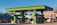 A vibrant green gas station with multiple fuel pumps is situated in a bright area surrounded by motorcycles, a van, and a distinct architectural building with intricate patterns. There are palm trees and flags in the background, adding to the lively and colorful ambiance.