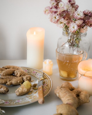 A cozy scene featuring a table with ginger roots arranged on a decorative plate alongside a knife. A steaming glass of herbal tea is nearby, with slices of ginger visible in the cup. Softly glowing candles provide a warm ambiance, while a jar of delicate pink flowers adds a touch of nature.