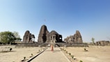An ancient stone temple with intricate carvings stands majestically in a spacious, open area. A single person in traditional attire is walking towards the temple, which has a prominent shikhara and a flag waving at its pinnacle. The surroundings include a few small trees and shrubs, suggesting a barren landscape with clear blue skies above.