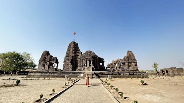 An ancient stone temple with intricate carvings stands majestically in a spacious, open area. A single person in traditional attire is walking towards the temple, which has a prominent shikhara and a flag waving at its pinnacle. The surroundings include a few small trees and shrubs, suggesting a barren landscape with clear blue skies above.