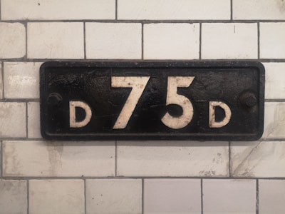 A rectangular, vintage sign mounted on a tiled wall displays the characters 'D 75 D' in white against a black background. The tiles are white with some wear and discoloration, lending an aged appearance to the scene.