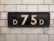 A rectangular, vintage sign mounted on a tiled wall displays the characters 'D 75 D' in white against a black background. The tiles are white with some wear and discoloration, lending an aged appearance to the scene.