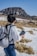 A person with long dark hair stands outdoors, holding a guidebook while wearing a white jacket and gray backpack. They are surrounded by a snowy landscape with patches of grass and a mountain in the distance under a clear blue sky.