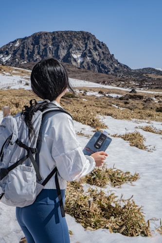 A person with long dark hair stands outdoors, holding a guidebook while wearing a white jacket and gray backpack. They are surrounded by a snowy landscape with patches of grass and a mountain in the distance under a clear blue sky.