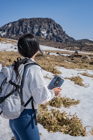 A person with long dark hair stands outdoors, holding a guidebook while wearing a white jacket and gray backpack. They are surrounded by a snowy landscape with patches of grass and a mountain in the distance under a clear blue sky.
