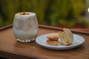 A glass filled with a creamy beverage, possibly a milkshake or a coffee drink with foam on top and sprinkled garnish, is placed on a wooden tray. Next to it, there is a white plate holding two pieces of a dessert that appears to be some sort of filled pastries or cookies with a creamy filling inside. The background is blurred with green foliage, suggesting an outdoor or garden setting.