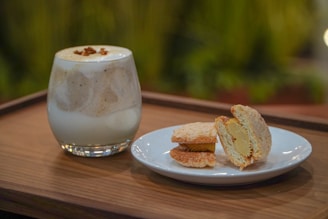 A glass filled with a creamy beverage, possibly a milkshake or a coffee drink with foam on top and sprinkled garnish, is placed on a wooden tray. Next to it, there is a white plate holding two pieces of a dessert that appears to be some sort of filled pastries or cookies with a creamy filling inside. The background is blurred with green foliage, suggesting an outdoor or garden setting.