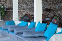 A cozy seating area with blue and gray cushions on a stone bench, set against a backdrop of a stone wall. Wooden tables with plants and small decor items are in the background, creating a relaxed atmosphere.