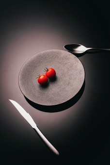 A minimalist setup with two small tomatoes placed on a textured gray plate set against a dark background. A shiny silver knife and spoon are positioned on either side of the plate, creating a balanced and elegant arrangement.