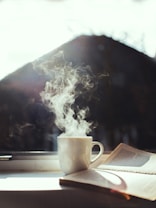 A steaming cup of coffee sitting on a windowsill beside an open book. Sunlight shines through the window, casting soft shadows.